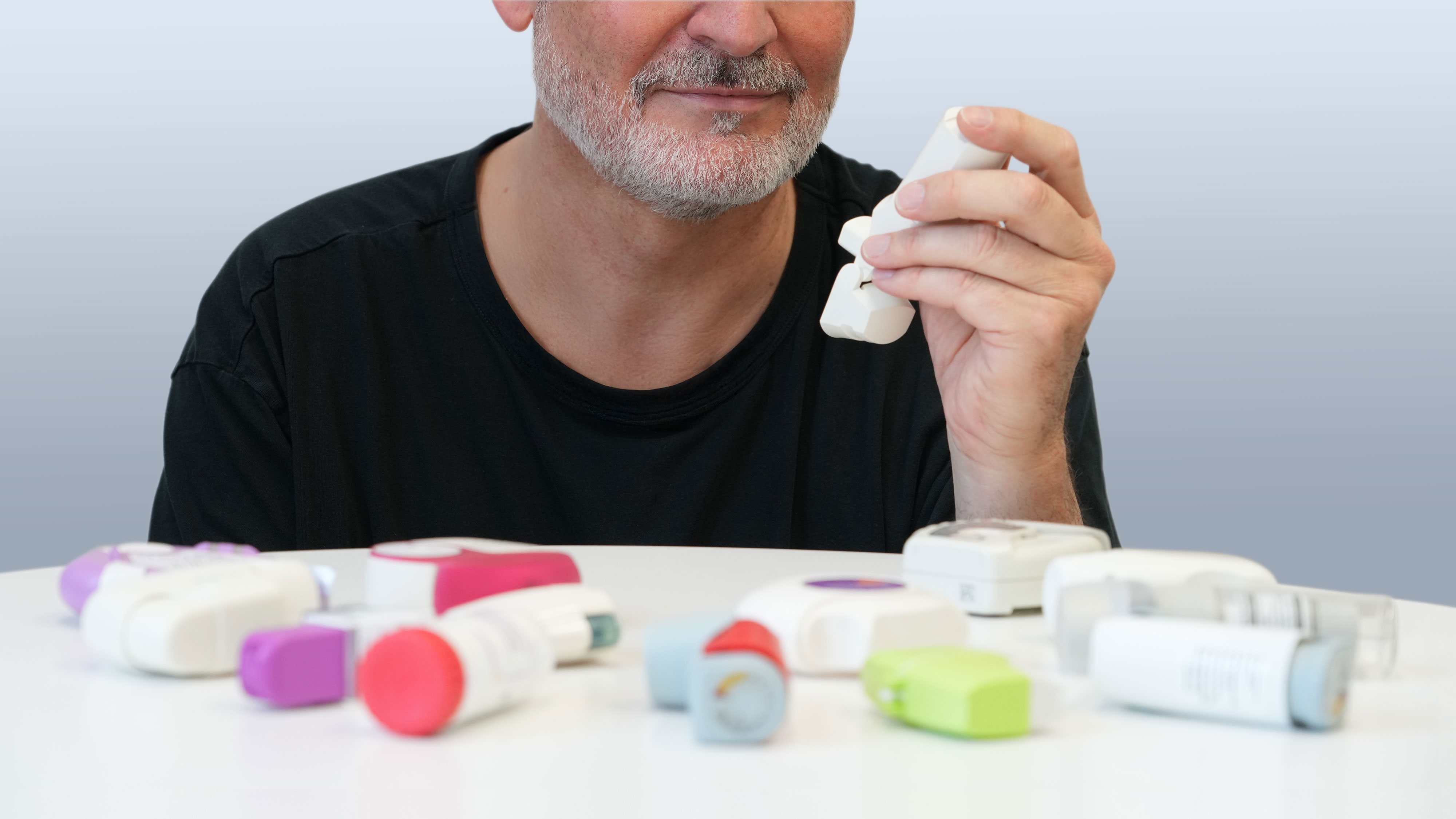 Männlicher Patient Mitte 50 beim Test verschiedener Inhalatoren für Asthma- und COPD-Patienten, die vor ihm auf einem Tisch ausgebreitet sind